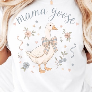 Puede incluir: Camiseta blanca con una ilustraci&oacute;n de un ganso con un lazo a cuadros, rodeado de flores y cintas. El texto "mama goose" est&aacute; escrito encima del ganso en una fuente de escritura. Un dise&ntilde;o encantador para una madre.