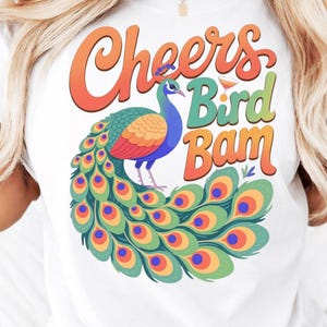 May include: White t-shirt featuring a colorful peacock illustration with the words "Cheers Bird Bam" in orange, green, and yellow. The peacock has blue, green, orange, and red feathers. A fun, whimsical design.
