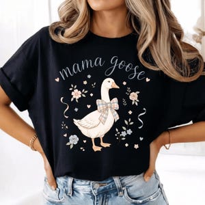 Puede incluir: Camiseta negra con la frase "mama goose" en cursiva. La camiseta presenta una ilustraci&oacute;n de un ganso con un lazo a cuadros, rodeado de motivos florales y cintas. Una parte superior informal y c&oacute;moda.