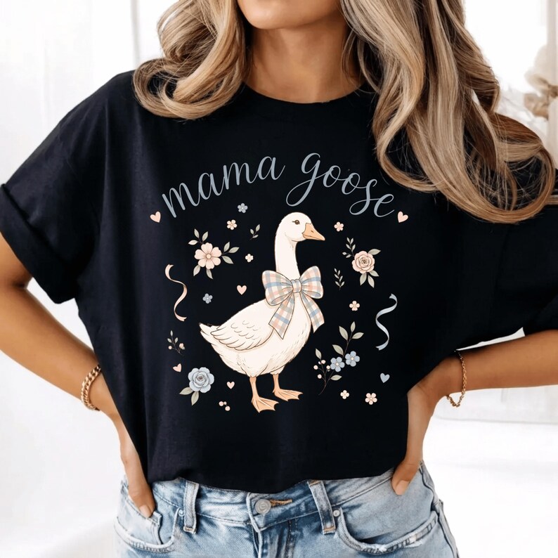 Puede incluir: Camiseta negra con la frase "mama goose" en cursiva. La camiseta presenta una ilustraci&oacute;n de un ganso con un lazo a cuadros, rodeado de motivos florales y cintas. Una parte superior informal y c&oacute;moda.