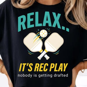 Könnte beinhalten: Schwarzes T-Shirt mit einem teal- und gelbfarbenen Pickleball-Motiv. Das Design enthält die Worte "RELAX...", zwei Schläger und einen Ball. Darunter steht "IT'S REC PLAY" und "nobody is getting drafted".