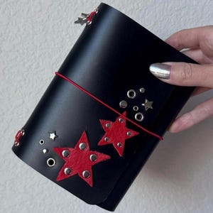 Op de afbeelding: Een zwarte leren journalomslag met twee rode leren sterapplicaties, zilverkleurige metalen studs en zilverkleurige stervormige klinknagels. De omslag wordt vastgezet met een dun rood elastisch koord en bevat diverse zilverkleurige metalen oogjes.