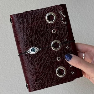 May include: A dark burgundy, textured leather-effect journal cover featuring a vertical row of silver-toned metal grommets and small rings. A thin black elastic cord wraps around the cover, secured by a silver-toned charm shaped like an eye.