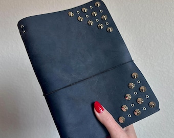 Puede incluir: Diario de cuero azul oscuro con tachuelas y ojales de metal plateado. El diario se mantiene cerrado con una banda elástica negra. La cubierta está decorada con múltiples tachuelas plateadas.