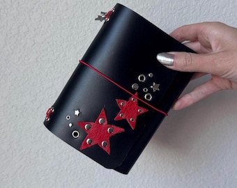 Puede incluir: Una cubierta de diario negra con dos grandes aplicaciones de estrellas rojas texturizadas y varios remaches metálicos plateados en forma de estrella y ojales circulares. Un fino cordón elástico rojo envuelve la cubierta con dijes de estrella.