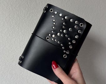 Puede incluir: Diario de cuero negro con tachuelas y ojales plateados. El diario se mantiene cerrado con una banda elástica negra. La cubierta está decorada con tachuelas de varios tamaños. Un accesorio elegante.