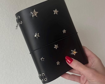 Puede incluir: Una funda de cuero negro para diario con tachuelas plateadas en forma de estrella dispersas por la parte frontal. La funda se asegura con una banda elástica negra fina e incluye ojales metálicos en el lateral para encuadernación.