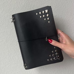 May include: A black leather journal cover featuring silver-toned star-shaped metal studs arranged in the top right and bottom right corners. A thin black elastic band wraps around the center, holding a small silver star charm.