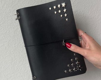 Puede incluir: Diario de cuero negro con adornos de estrellas plateadas. El diario se cierra con una banda elástica negra y presenta protectores de esquina de metal plateado. La cubierta está decorada con estrellas plateadas de varios tamaños.