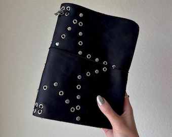 Puede incluir: Una funda de diario de cuero negro con una serie de ojales metálicos plateados dispuestos en un patrón de estrella en la parte frontal. La funda está asegurada con una banda elástica negra fina que envuelve horizontalmente el centro del objeto.