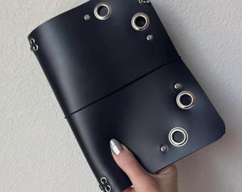 Puede incluir: Una funda de diario de cuero negro con ojales y remaches de metal plateado dispuestos en la superficie frontal. La funda está asegurada con una banda elástica negra fina envuelta horizontalmente alrededor del centro, con anillos metálicos.