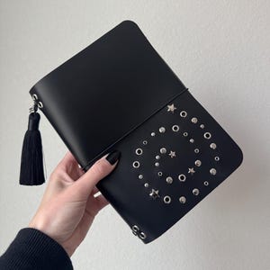 May include: Black clutch with silver-tone studs and star accents arranged in a spiral pattern. The clutch has a black tassel and a flap closure. The bag is held by a person.