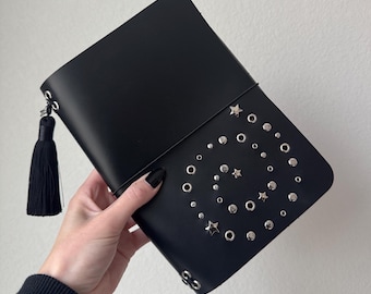 Puede incluir: Clutch negra con tachuelas plateadas y detalles de estrellas dispuestas en espiral. El clutch tiene una borla negra y un cierre de solapa. La bolsa es sostenida por una persona.