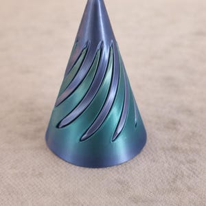 Könnte beinhalten: Ein kegelförmiges Deko-Objekt mit blau-grünem Farbverlauf. Das Objekt hat geschwungene, eingearbeitete Linien, die sich an den Seiten nach unten winden. Die Basis ist breiter als die Spitze, die spitz zuläuft.