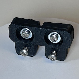 May include: A black plastic bracket with two silver screws and two silver bolts with washers. The bracket has a rectangular shape with two protruding sections. The screws and bolts are visible.