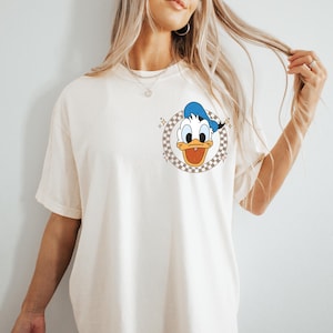 May include: Cream-colored t-shirt featuring a cartoon image of Donald Duck in a checkered circle. Donald Duck is wearing a blue sailor hat and has an open beak. The shirt has short sleeves.