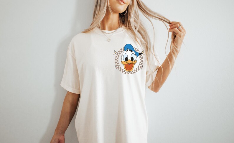 May include: Cream-colored t-shirt featuring a cartoon image of Donald Duck in a checkered circle. Donald Duck is wearing a blue sailor hat and has an open beak. The shirt has short sleeves.