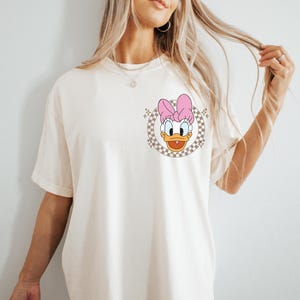 May include: Cream-colored t-shirt featuring a cartoon image of Daisy Duck with a pink bow. The graphic is set within a checkered circle. The shirt has short sleeves and a relaxed fit, suitable for casual wear.