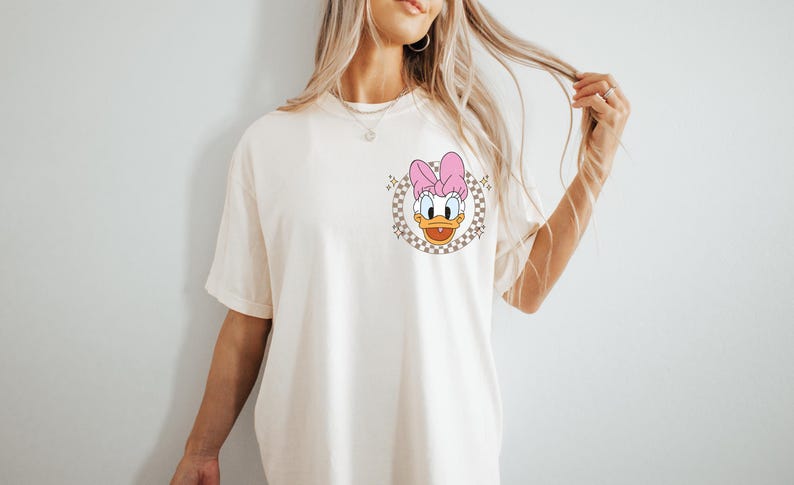 May include: Cream-colored t-shirt featuring a cartoon image of Daisy Duck with a pink bow. The graphic is set within a checkered circle. The shirt has short sleeves and a relaxed fit, suitable for casual wear.