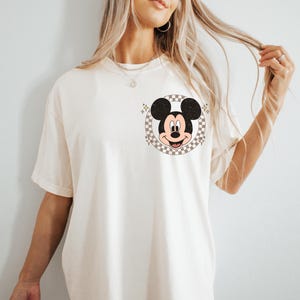 May include: Cream-colored t-shirt featuring a Mickey Mouse graphic. The graphic is a circular design with a checkered background and Mickey's face in the center. The shirt has short sleeves and a relaxed fit.
