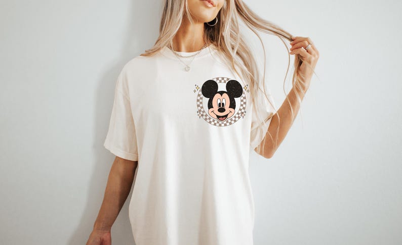 May include: Cream-colored t-shirt featuring a Mickey Mouse graphic. The graphic is a circular design with a checkered background and Mickey's face in the center. The shirt has short sleeves and a relaxed fit.