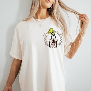 May include: Cream-colored t-shirt featuring a cartoon graphic of Goofy in a checkered circle. Goofy is wearing a green hat and has a large smile. The shirt has short sleeves and a relaxed fit.