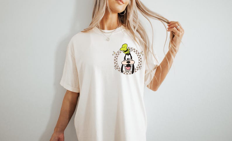 May include: Cream-colored t-shirt featuring a cartoon graphic of Goofy in a checkered circle. Goofy is wearing a green hat and has a large smile. The shirt has short sleeves and a relaxed fit.