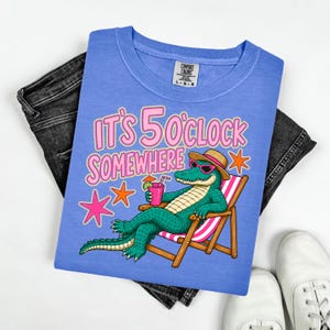 Puede incluir: Camiseta azul claro con el texto "IT'S 5 O'CLOCK SOMEWHERE" y un cocodrilo de dibujos animados con gafas de sol y sombrero, relajándose en una tumbona. La camiseta se muestra con vaqueros negros y zapatillas blancas.