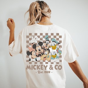May include: Cream-colored t-shirt featuring a retro-style graphic of Mickey Mouse, Minnie Mouse, Goofy, Donald Duck, and Daisy Duck. The design is set against a brown and white checkered background, with the text "Mickey & Co - Est. 1928".