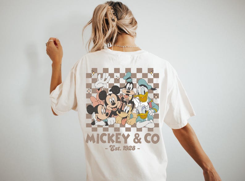May include: Cream-colored t-shirt featuring a retro-style graphic of Mickey Mouse, Minnie Mouse, Goofy, Donald Duck, and Daisy Duck. The design is set against a brown and white checkered background, with the text "Mickey & Co - Est. 1928".