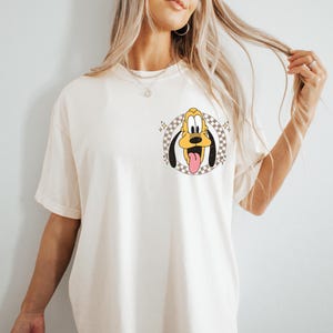 May include: Cream-colored t-shirt featuring a cartoon Pluto graphic. The graphic shows Pluto's face with his tongue sticking out, set within a checkered circle. The shirt has short sleeves and a relaxed fit.