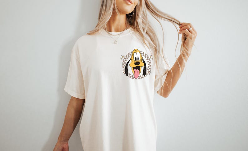 May include: Cream-colored t-shirt featuring a cartoon Pluto graphic. The graphic shows Pluto's face with his tongue sticking out, set within a checkered circle. The shirt has short sleeves and a relaxed fit.
