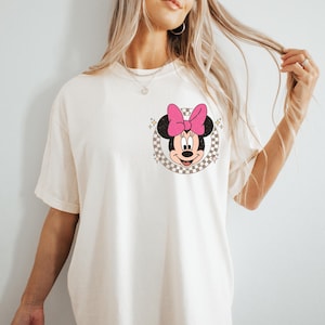 May include: Cream-colored t-shirt featuring a Minnie Mouse graphic. The graphic includes Minnie's face, a pink bow, and a checkered background. The shirt has short sleeves and a relaxed fit.