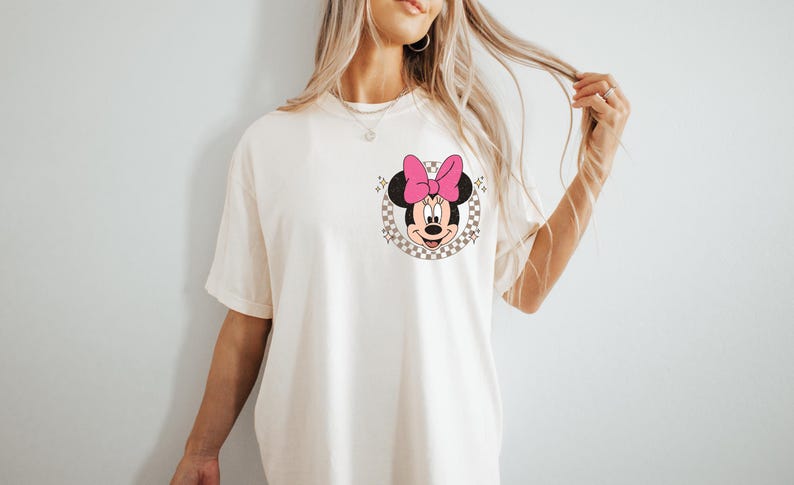 May include: Cream-colored t-shirt featuring a Minnie Mouse graphic. The graphic includes Minnie's face, a pink bow, and a checkered background. The shirt has short sleeves and a relaxed fit.