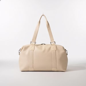 Peut inclure: Sac de sport crème avec deux poignées supérieures et une bandoulière amovible. Le sac a une forme rectangulaire avec une fermeture éclair et des accessoires en métal. Le sac est fait d'un matériau lisse semblable au cuir.