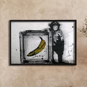 May include: A rectangular black frame containing a black and white print of a chimpanzee holding a smaller framed picture of a yellow banana. The artwork features a textured white background and includes the handwritten text Andy Warhol.