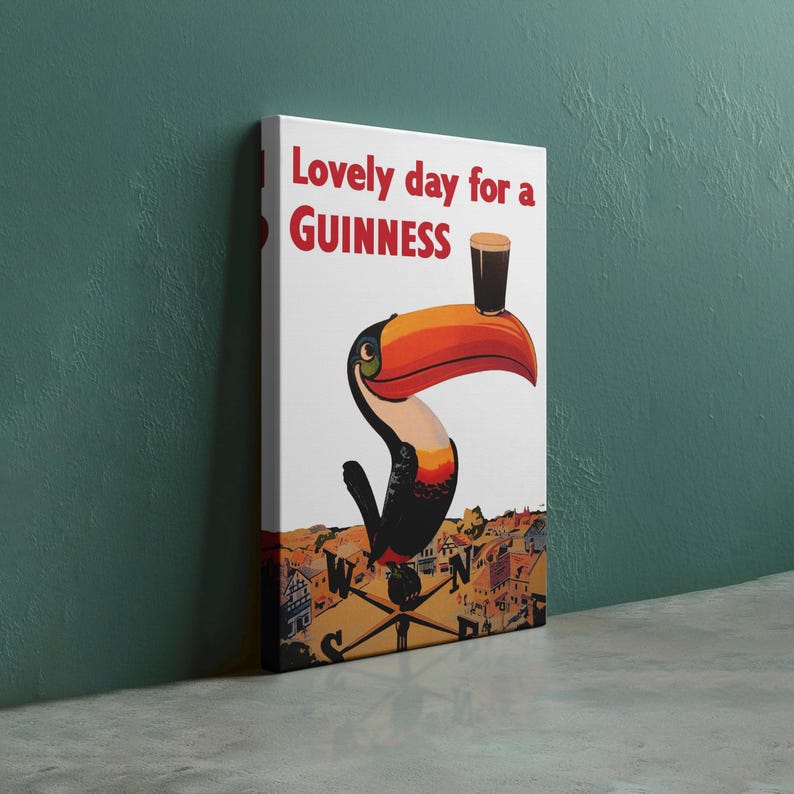 Op de afbeelding: Canvas print met een toekan die een pint Guinness vasthoudt. De tekst "Lovely day for a Guinness" is rood. De toekan heeft een grote oranje snavel. De achtergrond toont een stadsgezicht en een kompas.