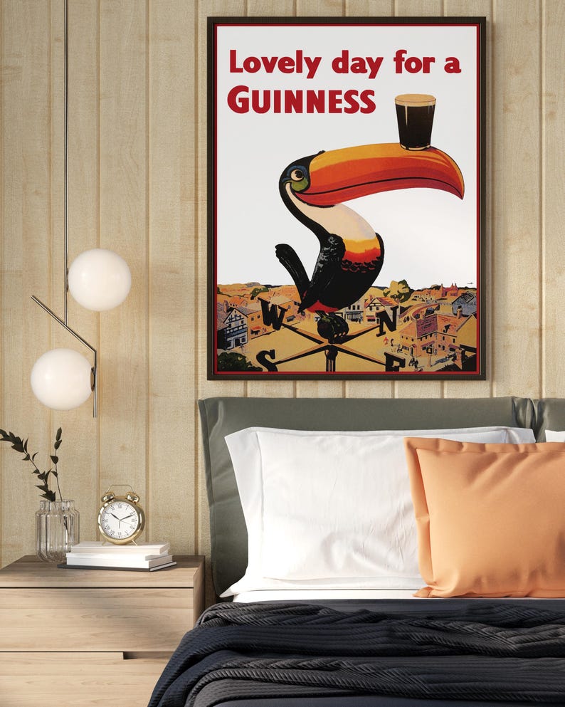May include: Framed Guinness advertisement featuring a toucan with a pint of stout on its beak. The poster reads "Lovely day for a Guinness" in red text. The artwork is set against a backdrop of a town and a compass.
