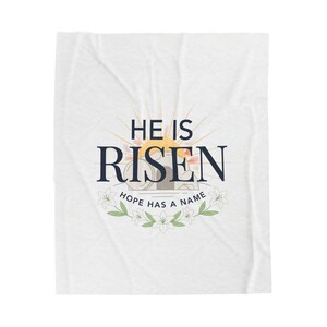May include: White blanket featuring the navy blue text "HE IS RISEN", with a sunburst design above and a floral wreath below. The words "HOPE HAS A NAME" are also present. The blanket has a subtle, textured pattern.