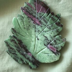 May include: A carved leaf-shaped stone with a green base color and dark purple and black veining. The stone is set against a light green fabric background. The leaf has detailed veining.
