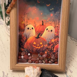 May include: A light wood frame containing a print of two white ghosts and orange pumpkins in a field, with loose red and orange stones at the bottom. A beaded bracelet hangs on the corner, and two raw mineral stones sit in front of the frame.