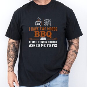 May include: Black t-shirt with the text "I HAVE TWO MOODS BBQ AND FIXING THINGS NOBODY ASKED ME TO FIX". The shirt has a barbecue grill and wrench graphic. The shirt is worn by a person.