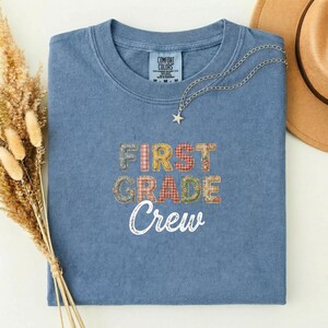 Peut inclure: Un t-shirt bleu Comfort Colors avec les mots "FIRST GRADE Crew" dans un motif patchwork. Un collier argenté avec un pendentif en forme d'étoile est drapé sur le t-shirt. Un chapeau beige est en arrière-plan.