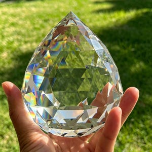 May include: A clear, faceted crystal prism in a teardrop shape. The crystal is held in a hand and reflects a rainbow of colours. The prism is displayed against a backdrop of green grass.