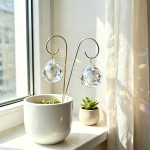 Könnte beinhalten: Zwei facettierte, klare Kristallkugeln hängen an geschwungenen Metallstäben, einer goldfarbenen und einer silberfarbenen. Die Stäbe stecken in einem weißen Keramiktopf, der eine kleine grüne Sukkulente enthält, wobei sich das Licht bricht.