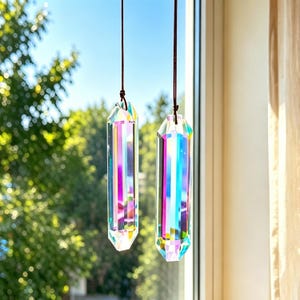 Könnte beinhalten: Zwei längliche, facettierte Kristallprismen mit einer lebendigen, irisierenden Oberfläche, die Regenbogenfarben reflektiert. Jedes Prisma ist vertikal an einer dünnen, dunkelbraunen Schnur aufgehängt und erzeugt so ein Lichtspiel vor einem Fenster.