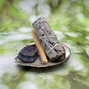 May include: A collection of spiritual items, including a white sage bundle tied with twine, a piece of palo santo wood, and a purple amethyst crystal, presented on an iridescent shell. The background is a soft green with bokeh.