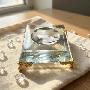 Pode incluir: Um suporte expositor retangular de vidro transparente com uma reentrância circular côncava no centro. O bloco de vidro é sustentado por quatro pés de metal dourado ornamentados com detalhes de volutas em cada canto.