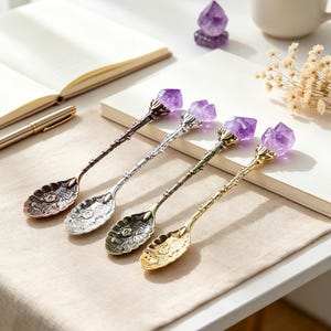 May include: Four metal spoons with ornate floral embossed bowls and textured stems, each topped with a raw purple amethyst crystal point. The set includes one copper, one silver, one antique bronze, and one gold-toned spoon, all arranged in a row.