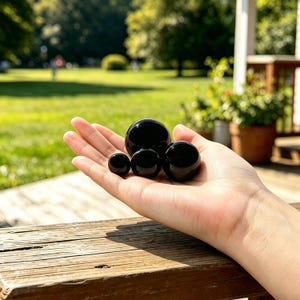 Op de afbeelding: Een hand die vier gepolijste zwarte bollen van verschillende grootte vasthoudt. De bollen hebben een glanzend, reflecterend oppervlak. De achtergrond toont een groen gazon en bomen.
